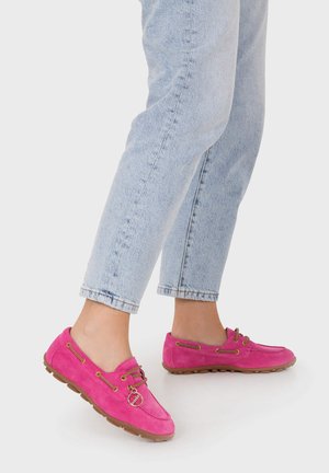 Osoba ubrana w jasnoniebieskie dżinsy i jasnoróżowe zamszowe mokasyny z brązowymi podeszwami i sznurówkami, stojąca na białym tle.