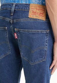 Jeans de mezclilla azul con un patrón de costura clara, que presentan un parche de cuero beige en la cinturilla trasera y dos bolsillos traseros con un diseño distintivo.