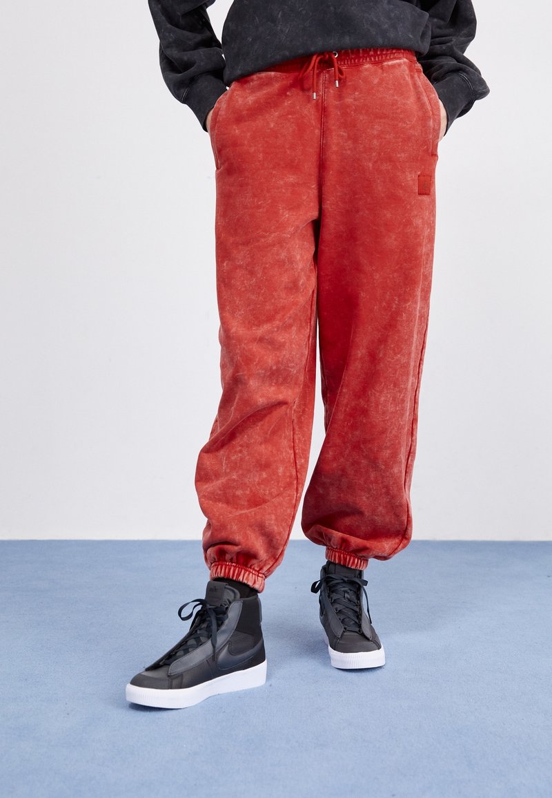 Rote, lässige Jogginghosen mit einer strukturierten, verblassten Optik und elastischem Bund. Kombiniert mit schwarzen High-Top Sneakers auf einem blauen Boden.