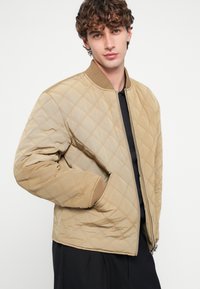 Prešívaná bombera bunda v béžovej farbe s rebrovaným golierom a manžetami, so zipsovým zapínaním a bočnými vreckami. Hladká, odolná textúra látky.