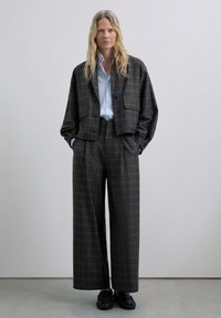 Chaquetón de cuadros cortos y pantalones de pierna ancha en tonos grises y marrones. Camisa azul claro por debajo. Zapatos negros completan el conjunto.