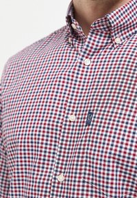 Chemise à carreaux avec un motif rouge, marine et blanc. Col court avec des boutons blancs et une étiquette bleue sur la poche poitrine.