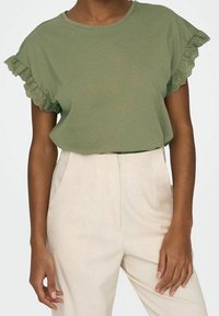 Olivgrön topp med dekorativ spetskant på korta ärmar, tillsammans med ljusa byxor med avslappnad passform och rund halsringning.