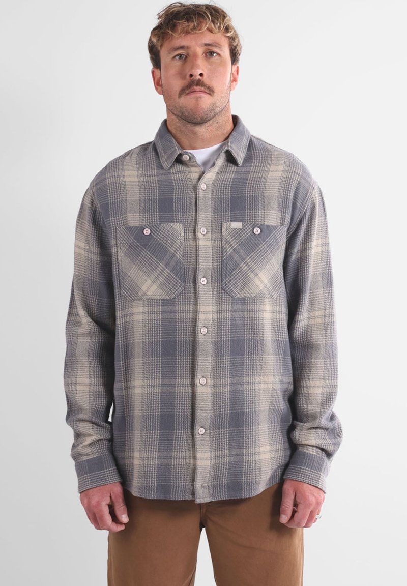 Homme de face portant une chemise à carreaux gris et beige avec deux poches poitrine et un pantalon marron sur fond uni.