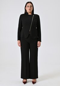 Zwarte op maat gemaakte blazer met een ritssluiting aan de zijkant, gecombineerd met zwarte wijde broek. Heeft een gestructureerd ontwerp en een gladde textuur.