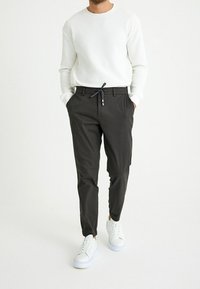 Leichtes weißes Langarmshirt mit geripptem Material, kombiniert mit schmal geschnittenen dunkelgrauen Hosen und weißen Sneakers mit blauer Sohle.