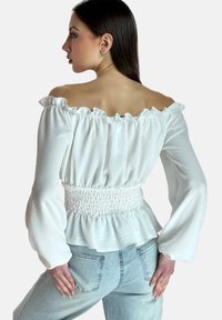 Weiße schulterfreie Bluse mit gerafftem Ausschnitt, geraffter Taille und langen Glockenärmeln. Getragen mit hellblauen Jeans.