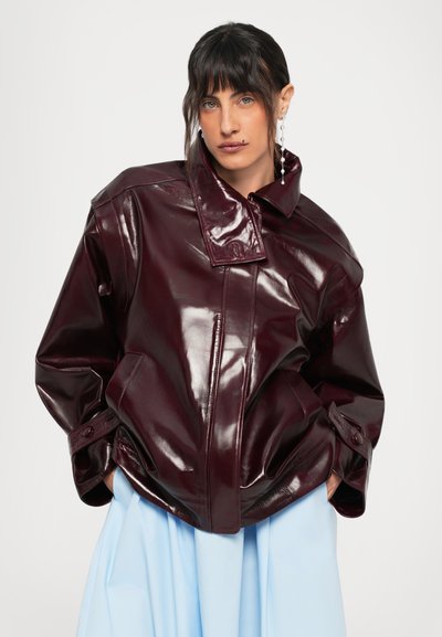 Persoană cu păr închis la culoare și breton, purtând o jachetă oversized din piele lăcuită de un burgund închis și pantaloni cu talie înaltă de un albastru deschis.