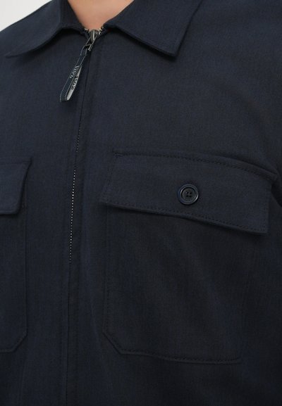 Navy blue zip-up shirt made of smooth fabric, featuring two buttoned chest pockets and a pointed collar with contrasting stitching.