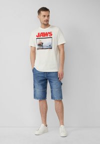 Biała bawełniana koszulka z czerwoną grafiką "JAWS" oraz nadrukiem zdjęcia. W połączeniu z niebieskimi szortami dżinsowymi i białymi trampkami. Prosty design.