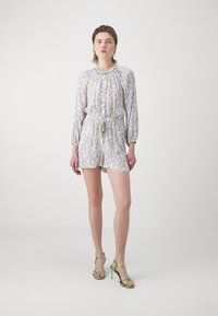 Ung kvinne står, iført en lys blomstret langermet romper med dusker, grønne sandal med høy hæl og gull hoops øredobber.