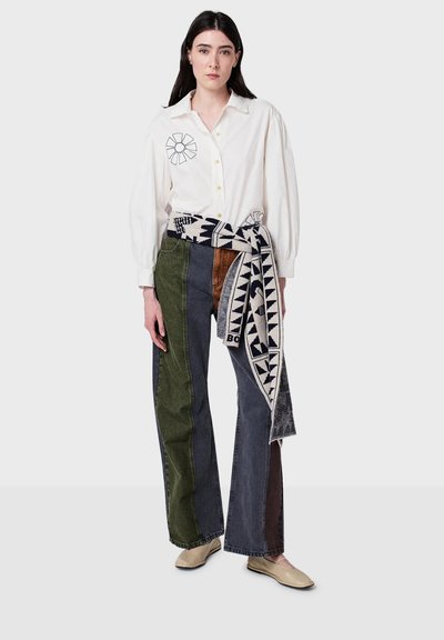 Camicia bianca in cotone con maniche a sbuffo e ricamo floreale, abbinata a jeans a patchwork multicolore con tonalità di verde, marrone e blu.