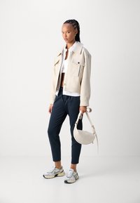 Beige cropped jack met twee zakken aan de voorkant, wit cropped shirt, marineblauwe taps toelopende broek en grijze sneakers. Houdt een ronde, crèmekleurige tas vast.