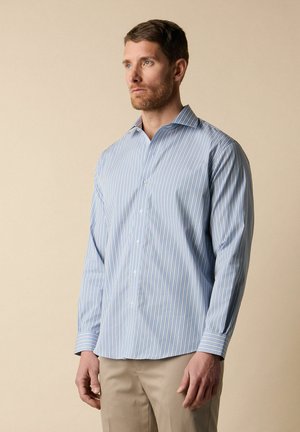 Mann trägt ein hellblau-weiß gestreiftes Hemd mit Knöpfen und beige Hose, steht vor einem schlichten beigen Hintergrund.