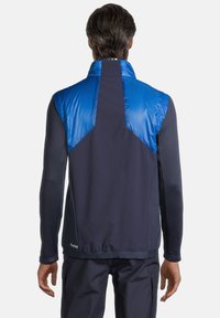 Veste hybride bleu et marine foncé présentant une section supérieure matelassée, une section inférieure douce, un col haut et un design sportif avec une coupe ajustée.