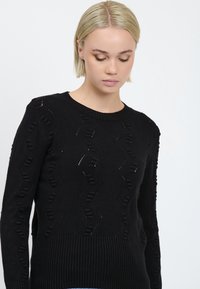 Maglione nero lavorato a maglia con un motivo testurizzato e dettagli a forare; colletto e orlo a coste. Maniche lunghe con un design intricato.