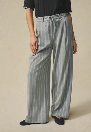 Pantaloni gri cu croială largă și dungi verticale, material texturat și talie legată. Purtați cu pantofi negri.