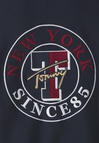 La tela azul marino presenta un logotipo bordado circular con "NUEVA YORK" en rojo, "T" en rojo y blanco, y "DESDE 85" en blanco, acentuado por una firma dorada.