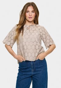 Blusa beige in pizzo con maniche scallop, scollatura tonda e dettaglio con fiocco. Abbinata a jeans in denim blu a vita alta.