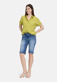 Groen linnen overhemd met knopen, blauwe denim bermuda shorts met rafelige zoom en witte slip-on sandalen. Model staat met handen op de heupen.