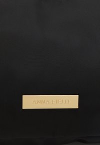 Superficie in tessuto nero con una piccola etichetta rettangolare dorata incisa con il nome "ANNA FIELD" centrato su di essa.