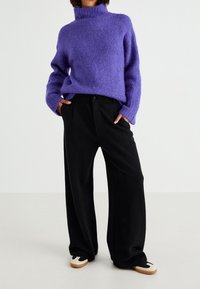Paarse, pluizige gebreide trui met een hoge halslijn, gecombineerd met zwarte broek met wijde pijpen. Schoeisel met een lichte zool en zwarte accenten.