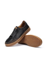 Sneaker in pelle nera con suola in gomma marrone; le caratteristiche includono un design perforato sulla punta, lacci e interno beige. Marchio sulla linguetta.