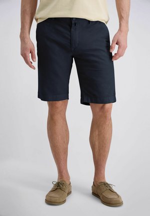 Man draagt marineblauwe op maat gemaakte korte broek met een beige overhemd en lichtbruine veterschoenen, staand op een effen achtergrond.