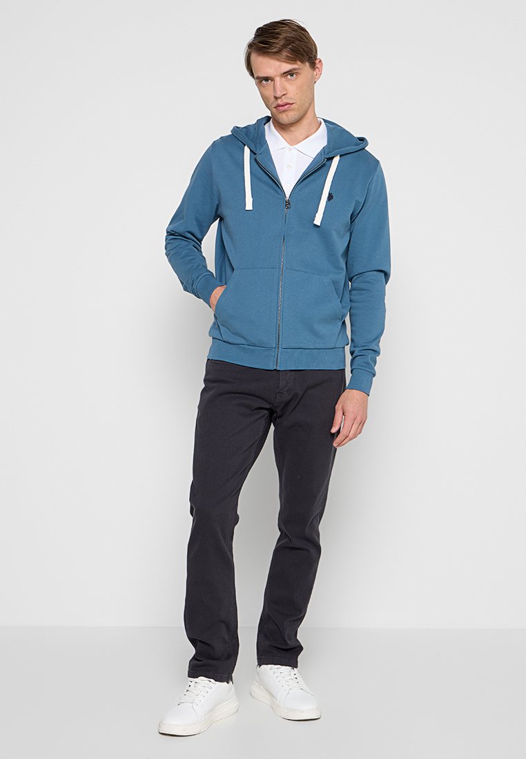 Mężczyzna w niebieskiej bluzie z kapturem na zamek, białej koszulce polo, czarnych spodniach i białych trampkach, stojący na tle jasnego tła.