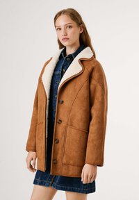 Giacca in ecopelle scamosciata marrone con colletto in shearling bianco, dotata di quattro bottoni, tasche laterali e una vestibilità comoda, indossata sopra un abito in denim.