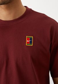 Burgundy puuvillapaita, jossa on pieni brodeerattu Nike-logo suorakulmaisen monivärisen kehyksen sisällä rinnassa. Lyhyet hihat, pyöreä kaula-aukko.