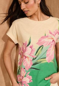 T-shirt floreale in morbido cotone, con fiori rosa pastello e foglie verdi su uno sfondo beige chiaro, maniche arrotolate e una vestibilità comoda.