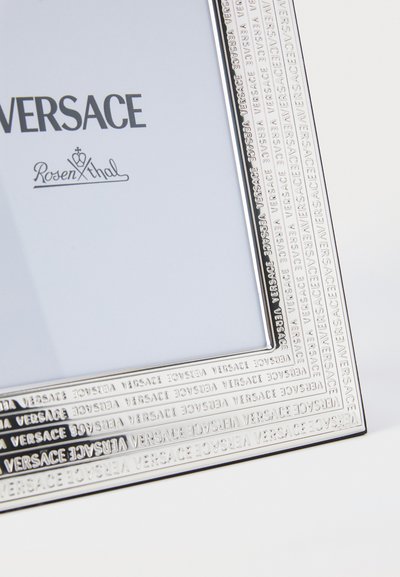 Silverrektangulär fotoram med präglad text "Versace" runt kanten. Slät yta, rymmer ett foto i storleken 4x6 tum.