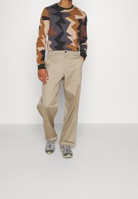 Koszula z długim rękawem w zygzakowy wzór w odcieniach brązu, czerni i beżu, zestawiona z jasnobeżowymi spodniami typu wide-leg i szarymi trampkami.