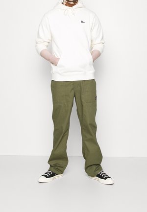 Person wearing a white hoodie, olive green pants, and black low-top sneakers standing against a plain light background.