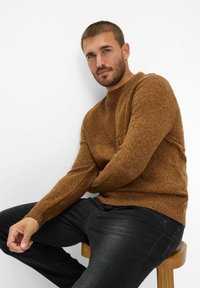 Bruin gebreid sweater met een patroon en ronde hals, gecombineerd met zwarte jeans. Zittend op een houten kruk tegen een witte achtergrond.