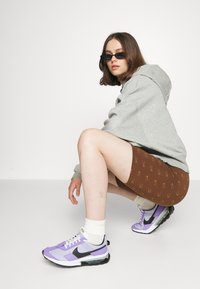 Sivi duksericu, smeđe kratke hlače s uzorkom, bele čarape i ljubičaste Nike patike s crnim akcentima. Model se čuči na beloj pozadini.