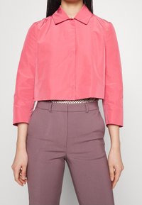 Kort jas in felroze, met een klassieke kraag en lange mouwen, gemaakt van gladde stof. Gedragen over een broek met hoge taille.