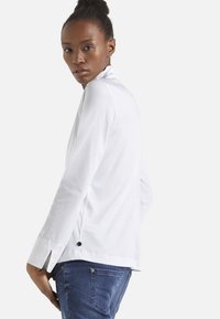 Camicia bianca a maniche lunghe con fessure laterali, texture sottile e dettagli in bottone neri, abbinata a jeans in denim blu.