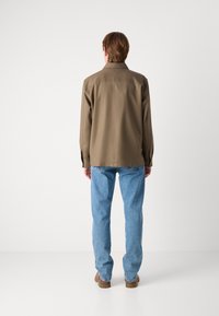 Camisa marrón de manga larga con cuello, de corte relajado, combinada con jeans azul claro. La camisa tiene una textura suave y un detalle mínimo.