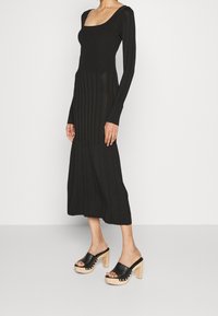 Donna che indossa un abito midi a maniche lunghe nero e tacchi a piattaforma neri con punta aperta e tacco in legno, in piedi su uno sfondo uniforme.