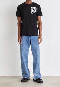 Černé bavlněné tričko s bílým grafickým potiskem na hrudi, doplněné světle modrými širokými džínami a černými teniskami.