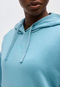 Κοντινό πλάνο ενός ατόμου που φοράει ένα ανοιχτό μπλε φούτερ με κορδόνια και ένα μικρό κεντητό λογότυπο Puma στο στήθος.