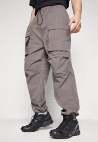 Šedé cargo kalhoty vyrobené z lehkého materiálu, které mají několik bočních kapes, elastický pas a stažené kotníky. Spárované se černými botami.