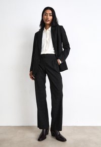 Costume noir à rayures avec une veste sur mesure et un pantalon droit, associé à un chemisier blanc et des bottes à talons foncées. Texture de tissu lisse.