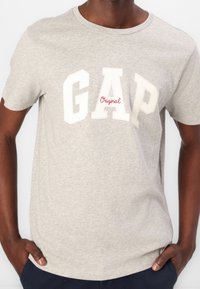 Mand iført en lys grå GAP T-shirt med "Original" i rød skrifttype, hænderne i lommerne, står mod en ensfarvet baggrund.