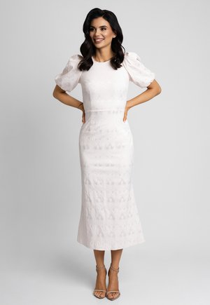 Kobieta o ciemnych włosach, ubrana w białą, teksturowaną sukienkę midi z bufiastymi rękawami i beżowe szpilki z paskiem na kostce, stojąca z rękami na biodrach.