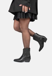 Botas de vaquero de cuero negro con punta afilada y tacones bajos, que presentan patrones decorativos de costura en los lados y altura hasta el tobillo.