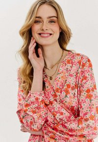 Kvetinová blúzka v ružovej farbe s červenými a oranžovými kvetmi, dlhé rukávy s priliehajúcimi manžetami, výstrih do V, skombinovaná s vrstvenými zlatými a perlovými náhrdelníkmi.
