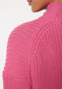 Pull rose tricoté avec un col montant, présentant un motif texturé côtelé sur les épaules et les manches. Fil doux et épais.